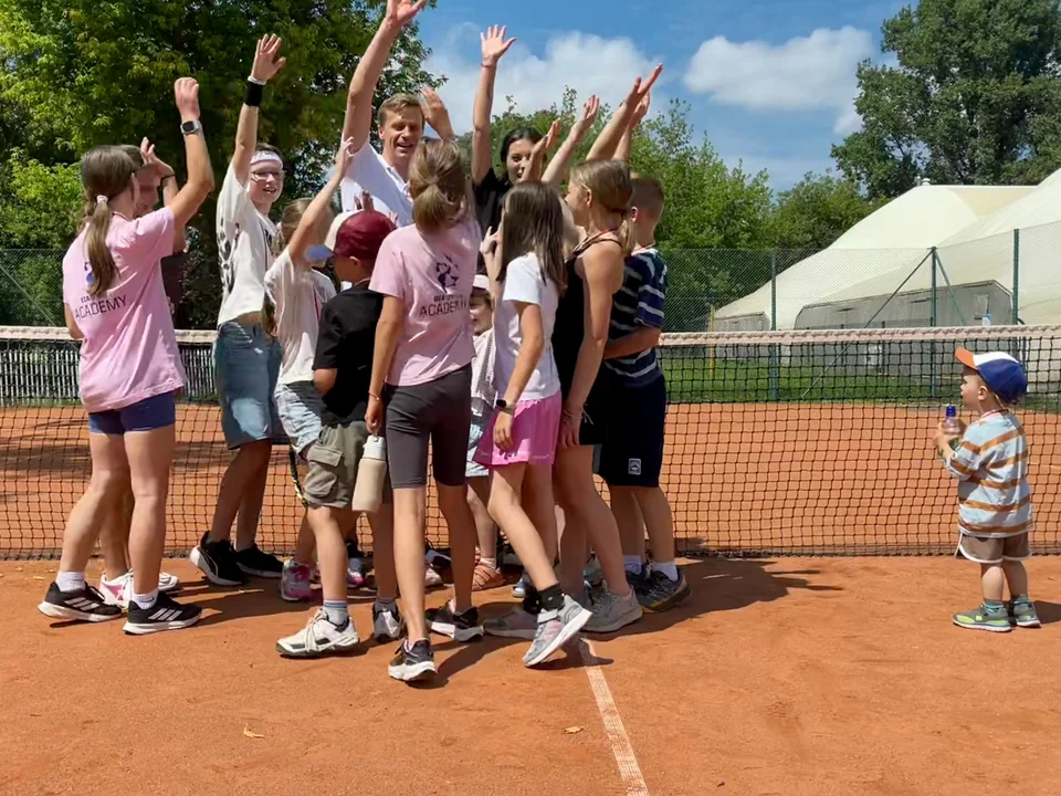 Dzieci i dorośli unoszą ręce przy siatce tenisowej